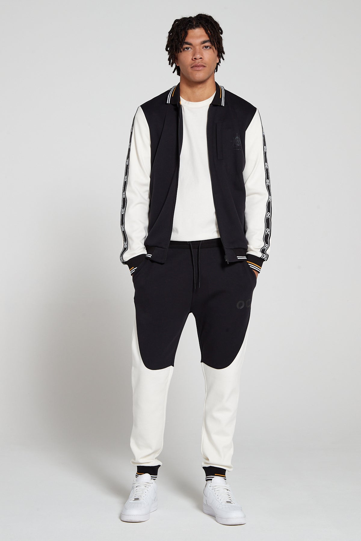 Full look of a model wearing oversized Original Creator Zip Jacket in black and white color. Model has the hoodie zipper opened and has hands in jogger pockets. Model is showing off complete Track set with white and black joggers and white reflection T shirt for the full look