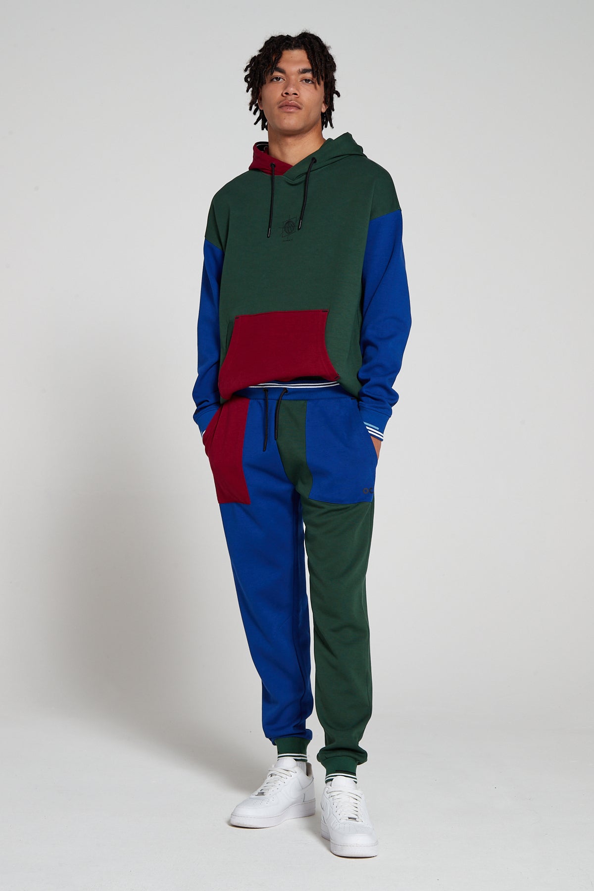 Full look of a model wearing Original Creator Classic in Multi color pattern with one leg in green and the other leg in blue color. Model is highlighting the combination with a multi color hoodie to complete the track set