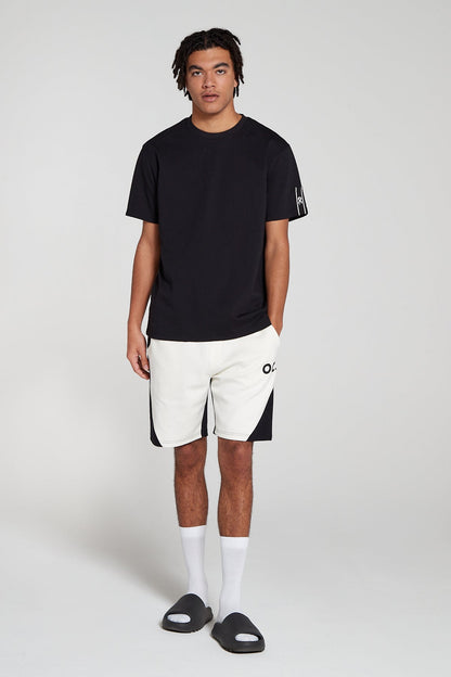Full look of a model wearing Original Creator Track 2.0 Retro Zip Pocket Shorts in Jet black Color with OC branding on left thigh and wearing a complementing black reflection T shirt with one hand in the shorts pocket.