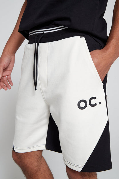 A close up side shot of Original Creator Track 2.0 Retro Zip Pocket Shorts in Jet black Color. Model is showcasing the OC branding on left thigh and the color combination of black and white in the shorts. Model has his hands in the shorts pockets and has the shirt tucked in from front