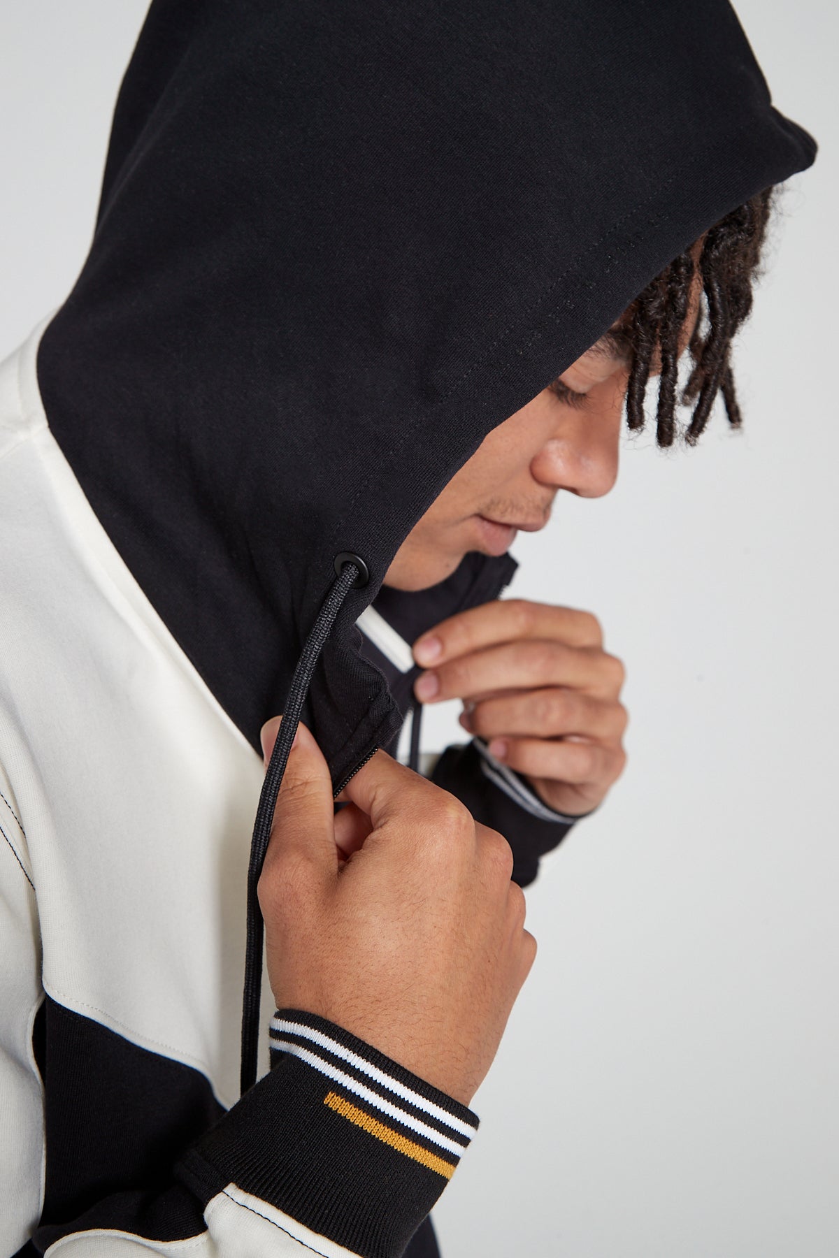 Close up sideview model wearing an Original Creator Retro Zip Hoodie in Jet black color. Showing off the hoodie with openned zip and comfortably wearing the hood. Model is also showing off the unique white and yellow lining on wrists while holding the zipper tape