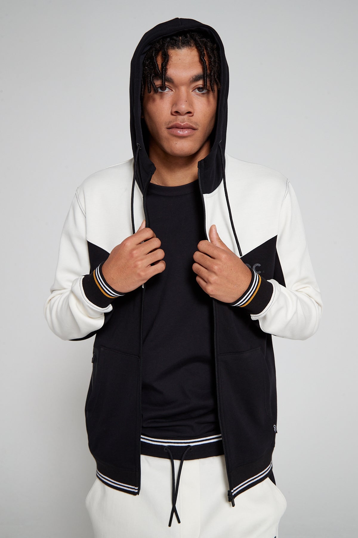 Front of a model wearing an Original Creator Retro Zip Hoodie in Jet black color. Showing off the hoodie with openned zip and comfortably wearing the hood. Model is also wearing Black reflection T shirt underneath to complete the track set