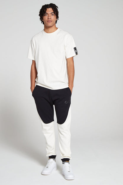 Full look of a model wearing Original Creator Reflection T-Shirt in Off white color with minimum branding on left sleeve and features water repellent technology. Model is also wearing Original creator Retro joggers in black and white color 