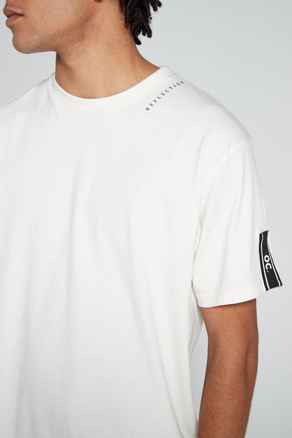 Model looking away showing close up view wearing Original Creator Reflection T-Shirt in Off White color with reflection written near the round neck and features water repellent technology and brand taping on the left sleeve