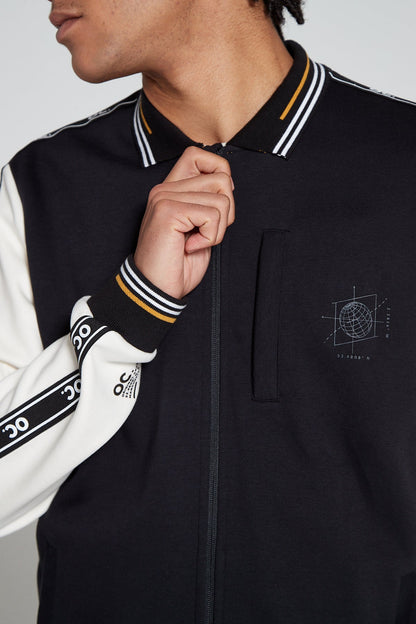 Front close up view of a model looking away wearing an Original Creator Zip Jacket in black and white color. Model is closing the zipper and Showing off the OC signature taping on the sleeves and the white and yellow lining on the wrist. Product feaures globe branding on the left zip pocket on chest 