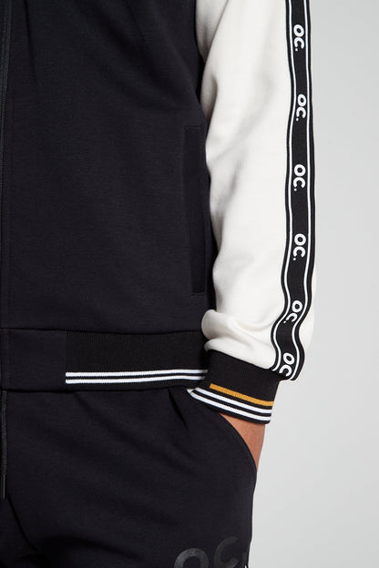 Close up bottom right view of Original Creator Zip Jacket in black and white color highlighting OC signature taping on left sleeve. Model is showing off the unique yellow and white on wrists of Jacket