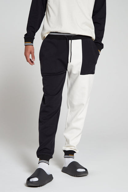A bottom half shot of model wearing Original Creator Track 2.0 Classic Joggers in Jet black Color with one leg in jet black and the other leg in off white white color. Model is highlighting the combination with long sleeve off white T shirt with black sleeves to match with white and black joggers style.