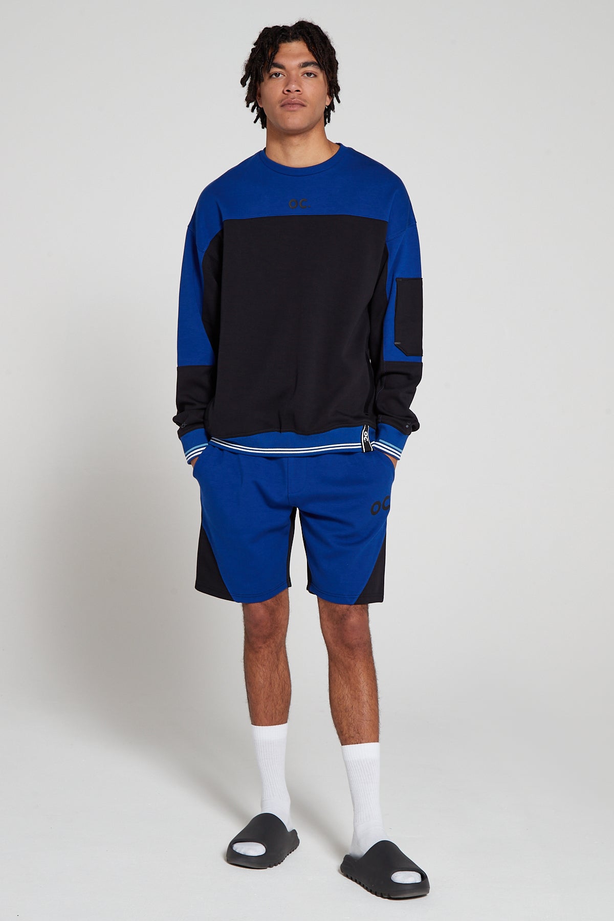 Full look of a model wearing Original Creator Track 2.0 Retro zip pocket Shorts in Olympic Blue color with OC branding on left thigh and wearing a matching black and blue zip pocket sweatshirt with both hands in the shorts pockets.
