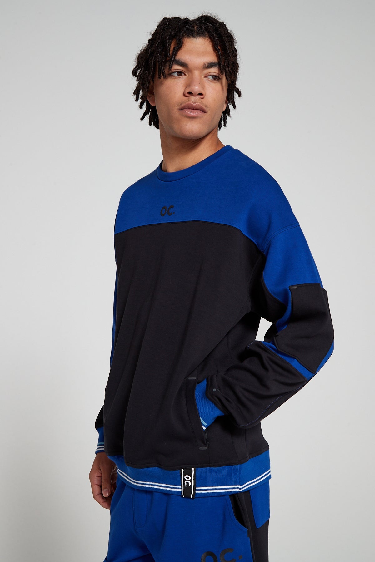 Model showing side view wearing Oversized Original Creator Retro Zip pocket Sweatshirt in Olympic Blue color highlighting zip pocket on sleeve and is putting hand in the side pocket. Showing off the complete set with color combination of black and blue on sweatshirt with and blue and black retro shorts.