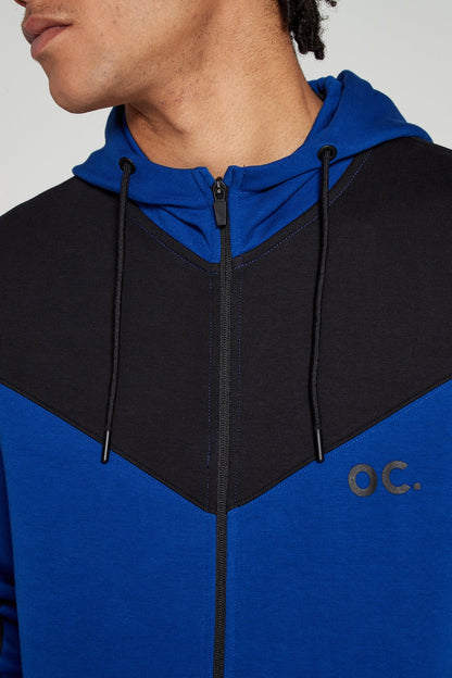 Close up center view of Original Creator Retro Zip Hoodie in Olympic Blue color. Model Showing off the hoodie with closed zip along with minimal OC branding on upper left side of chest. Model is looking away showing off the unique black and blue color combination of hoodie