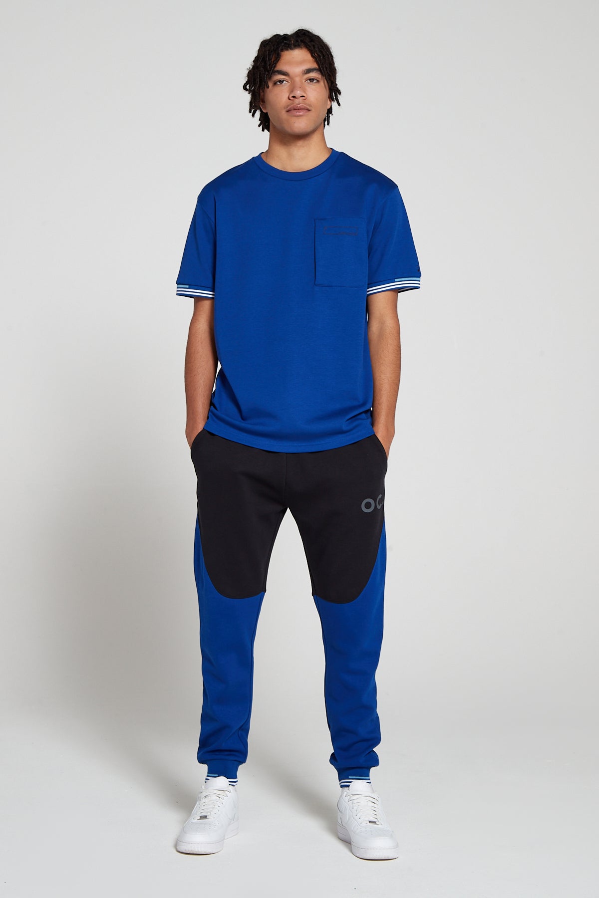 Full look of a model wearing Original Creator Retro Joggers in Olympic Blue color featuring a blue zip pocket T shirt and putting hands in the Jogger pockets. The joggers are in black and blue color combination. 