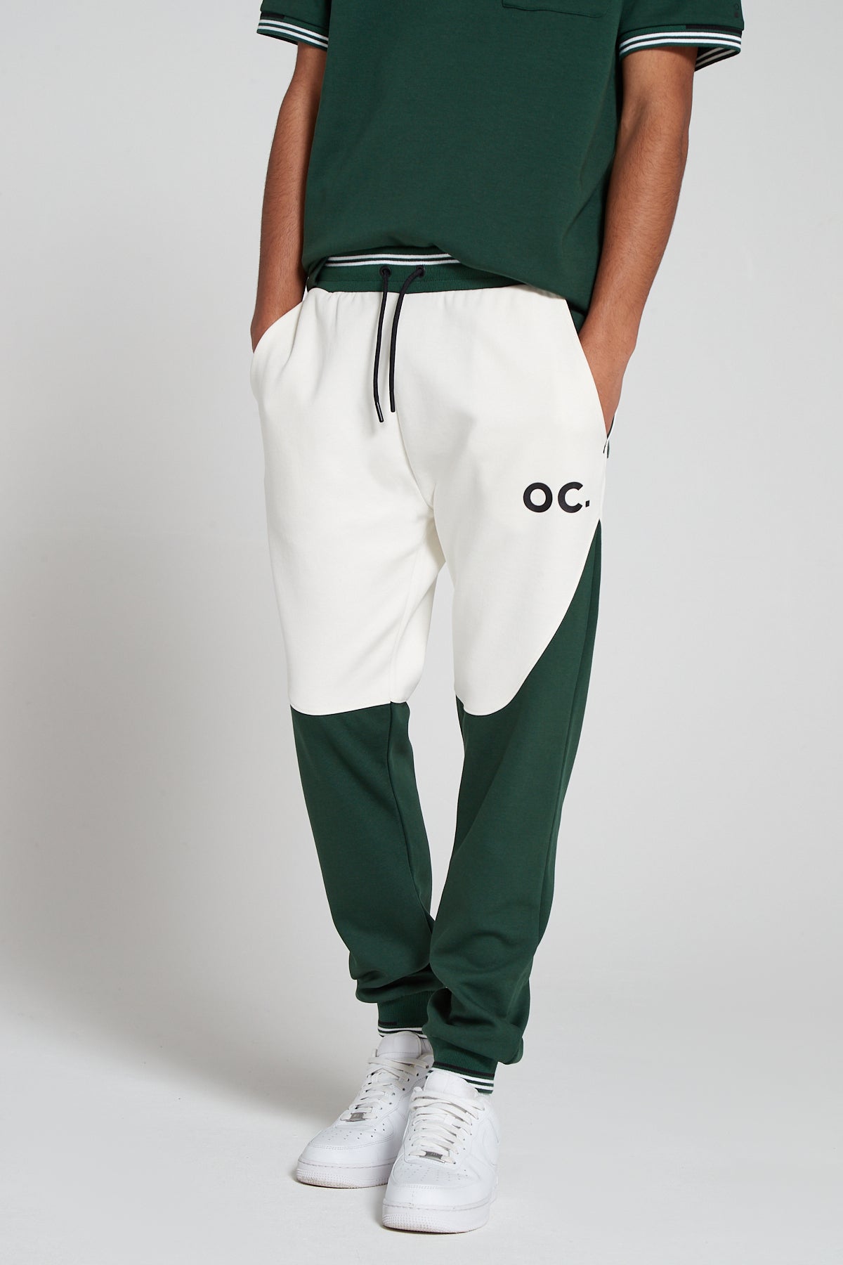 A bottom half shot of model wearing Original Creator Track 2.0 Retro Joggers in Varsity Green Color. Model is showcasing the length of Joggers and branding on the left thigh. Also highlighting the color combination of green and white on joggers.