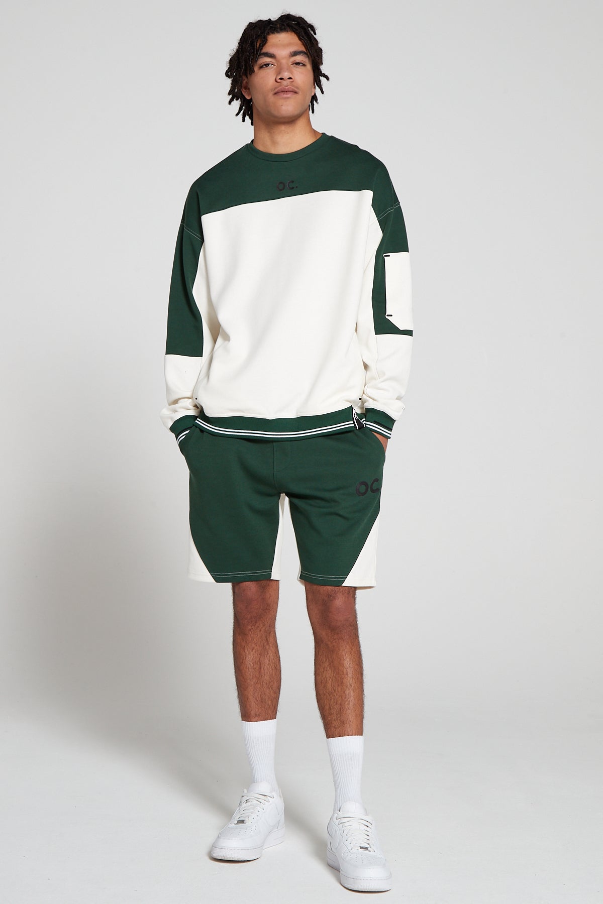 Full look front view of a model wearing Original Creator Track 2.0 Retro zip pocket Shorts in Varsity Green color with OC branding on left thigh and wearing a matching Green and White zip pocket sweatshirt with both hands in the shorts pockets.