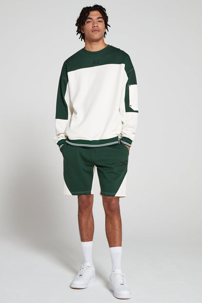 Full look front view of a model wearing Original Creator Track 2.0 Retro zip pocket Shorts in Varsity Green color with OC branding on left thigh and wearing a matching Green and White zip pocket sweatshirt with both hands in the shorts pockets.