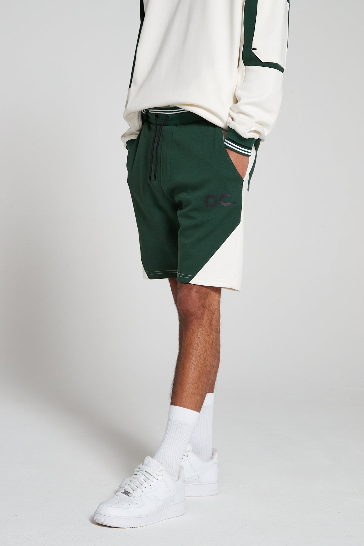 Side view of a model wearing Original Creator Track 2.0 Retro zip pocket Shorts in Varsity Green color and wearing a complementing green and white sweatshirt with both hands in the shorts pockets. Product features OC branding on the left thigh