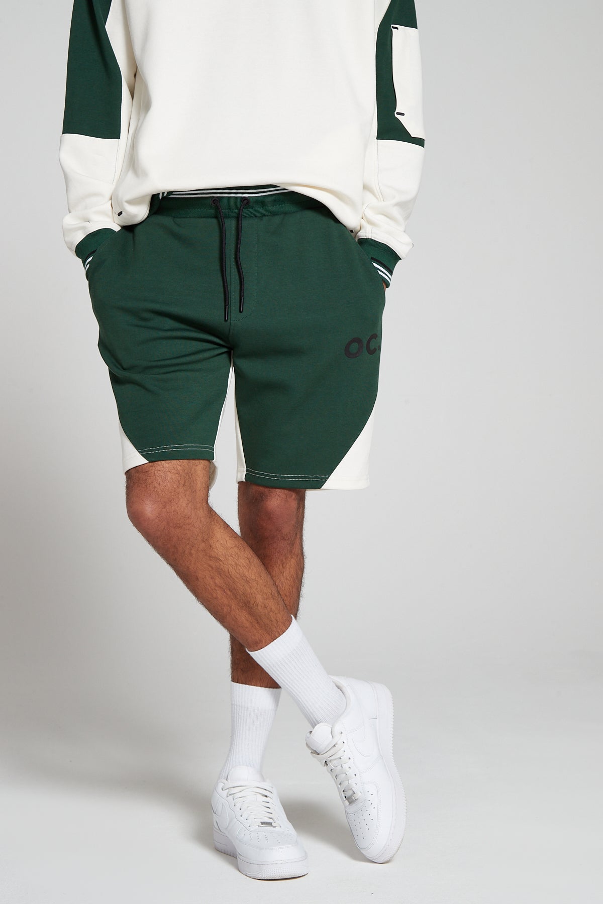 A bottom half side shot of model posing in crossed legs wearing Original Creator Track 2.0 Retro zip pocket Shorts in Varsity Green color. Model is showcasing the length of shorts and has hands in the pockets. Also highlighting the OC branding on right thigh.