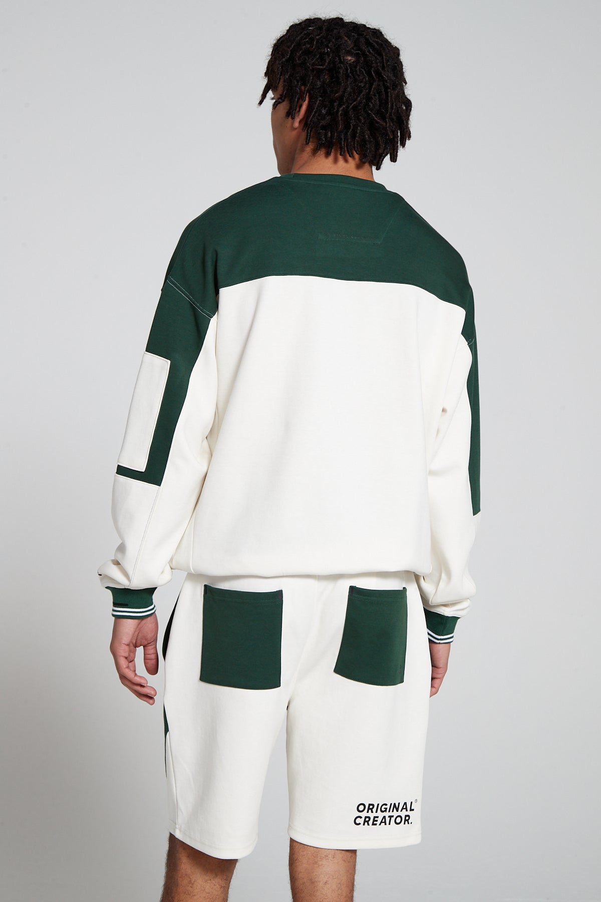 Full look back view of a model wearing Original Creator Track 2.0 Retro zip pocket Shorts in Varsity Green color with Horizontal Original Creator Branding on bottom right corner of shorts. The model is wearing a matching Green and White zip pocket sweatshirt showcasing the combination of green and white.