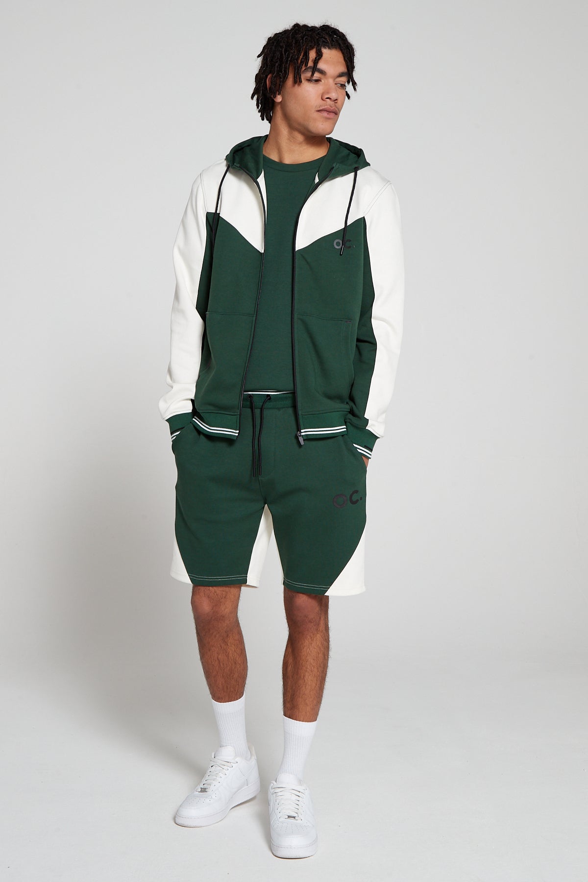 Full look of a model wearing oversized Original Creator Retro Zip Hoodie in Varsity Green color. Model has the hoodie zipper opened and has hands in shorts pockets. Model is showing off complete Track set with Varsity Green Retro shorts and Varsity Green zip pocket T shirt for the full look