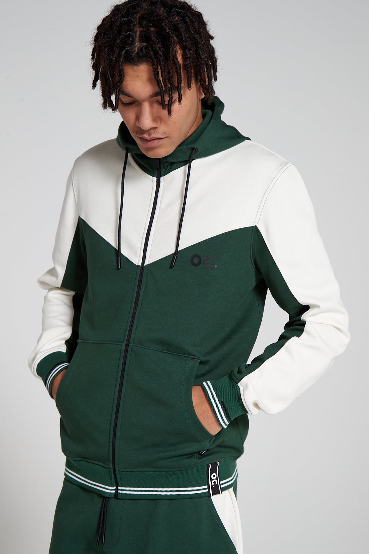 Front view of a model lookling down wearing an Original Creator Retro Zip Hoodie in varsity green color. Showing off the hoodie with closed zip and the color combination of white with green on hoodie. Hoodie features minimal OC branding on upper left side of chest with 4 pockets on the hoodie