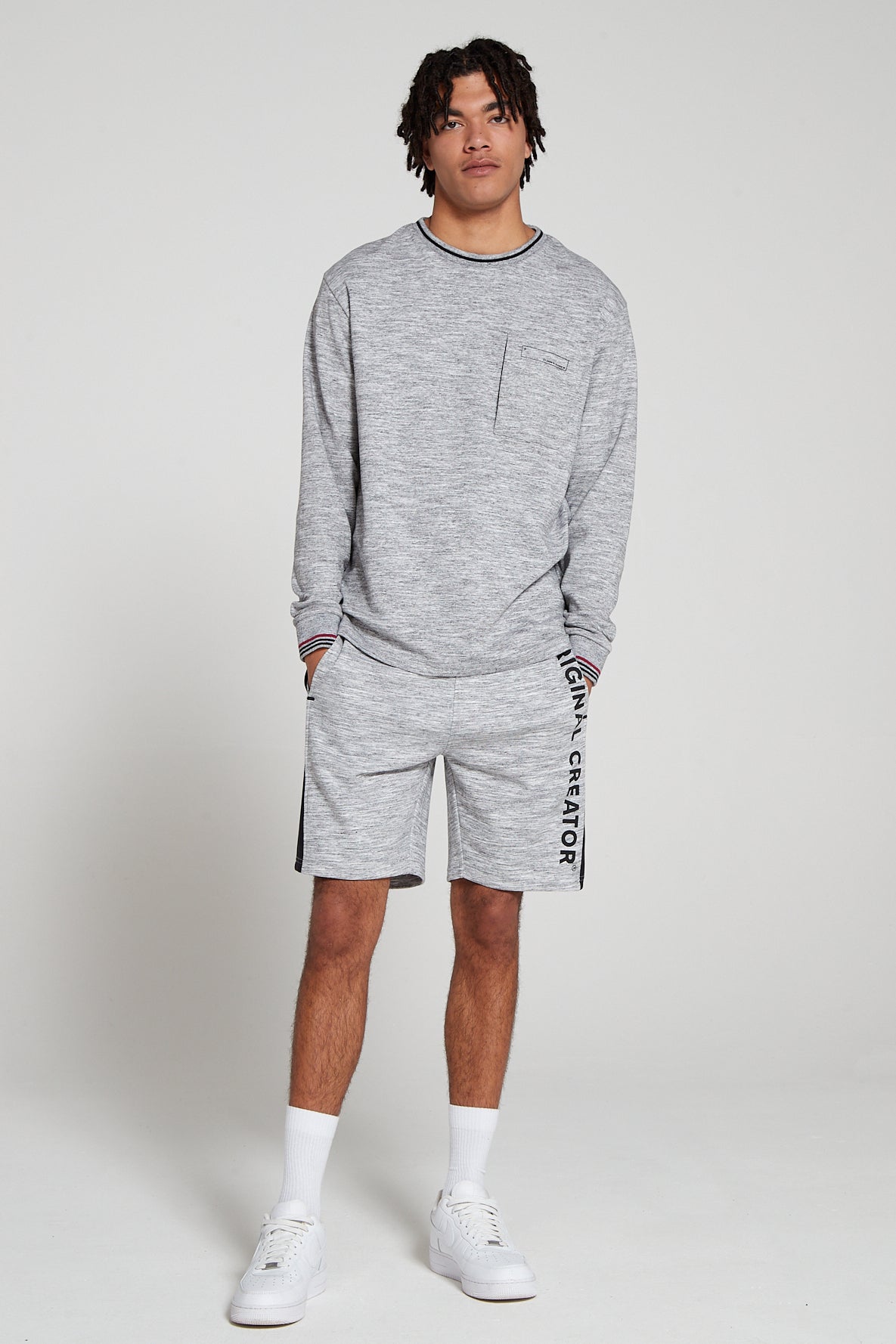 Full look of a model wearing Original Creator Granite Grey zip pocket shorts with vertical branding on left thigh and featuring a matching Track set of long sleeve granite grey T shirt and putting hands in the shorts pockets