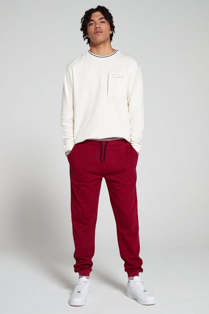 Full look of a model wearing Original Creator Classic Cranberry Red Joggers featuring a long sleeve Off white T shirt and putting hands in the Jogger pockets. The joggers are in red color. 