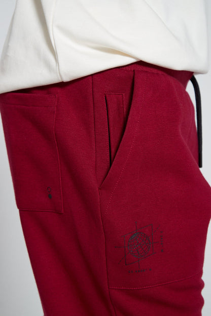 A side angle and close up shot of Original Creator Cranberry Red Classic Joggers with a total of four side pockets includes two zip pockets. Close up shot also highlights a back pocket and globe branding under the pocket on right side.