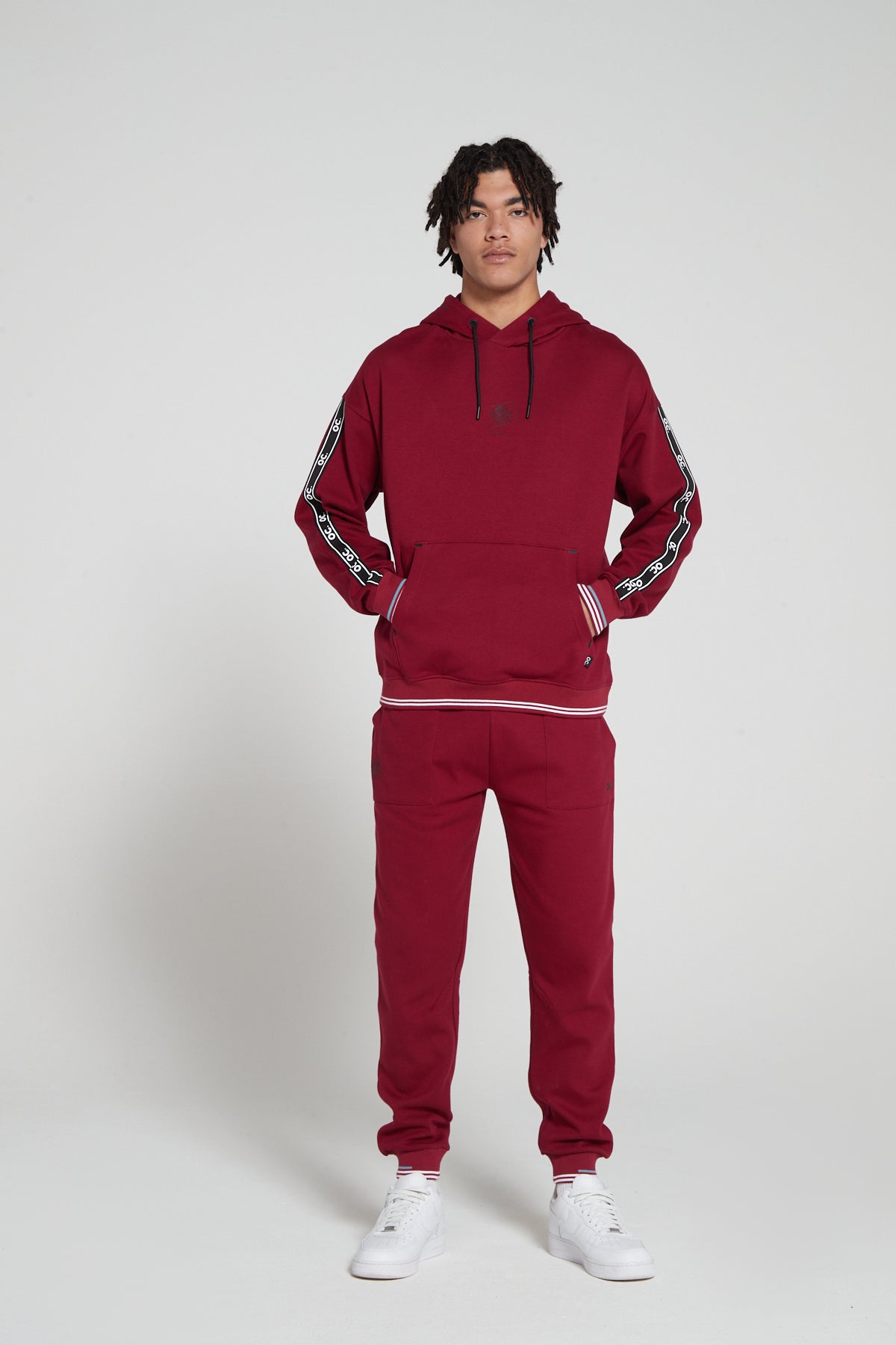 Full look of a model wearing oversized Original Creator  Classic Hoodie in Cranberry Red color with OC signature taping on both sleeves and features water repellent technology zip pocket along a matching Track set of Original creator classic Cranberry Red Joggers and hand in the kangaroo pocket of hoodie