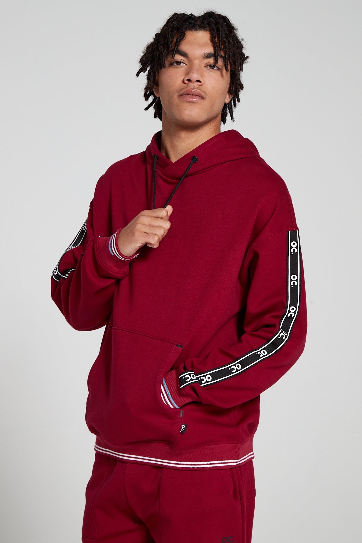 Model is tugging down the drawstrings of an Original Creator oversized Classic Hoodie in Cranberry Red color looking Straight. Showing off signature OC taping on both sleeves and globe print on center chest. 