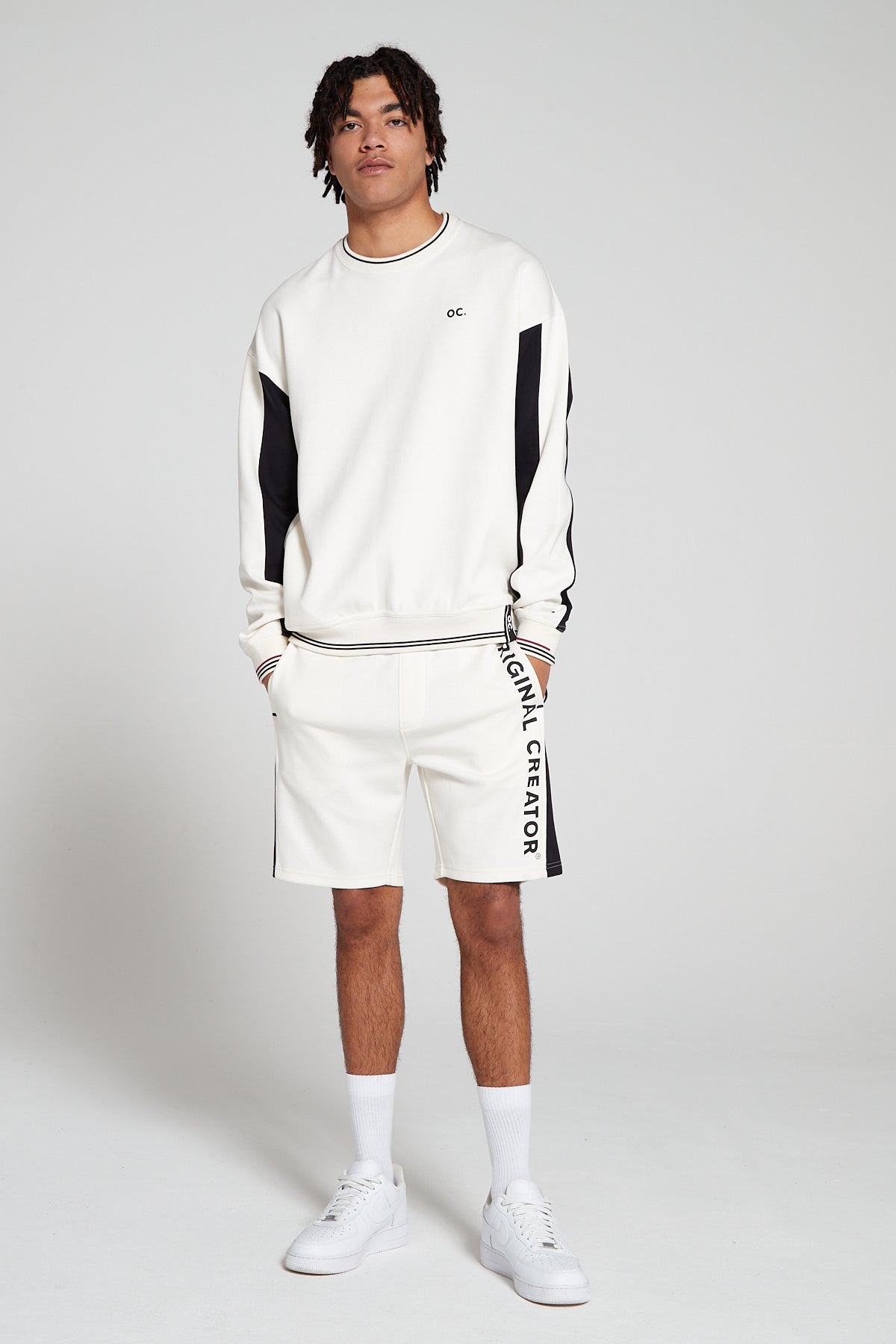 Full look of a model wearing oversized Original Creator Retro Sweatshirt in Off white color. Minimal branding and color combination of black and grey on sweatshirt. Showing off the complete set with Retro Off white shorts with hands in the shorts pockets.