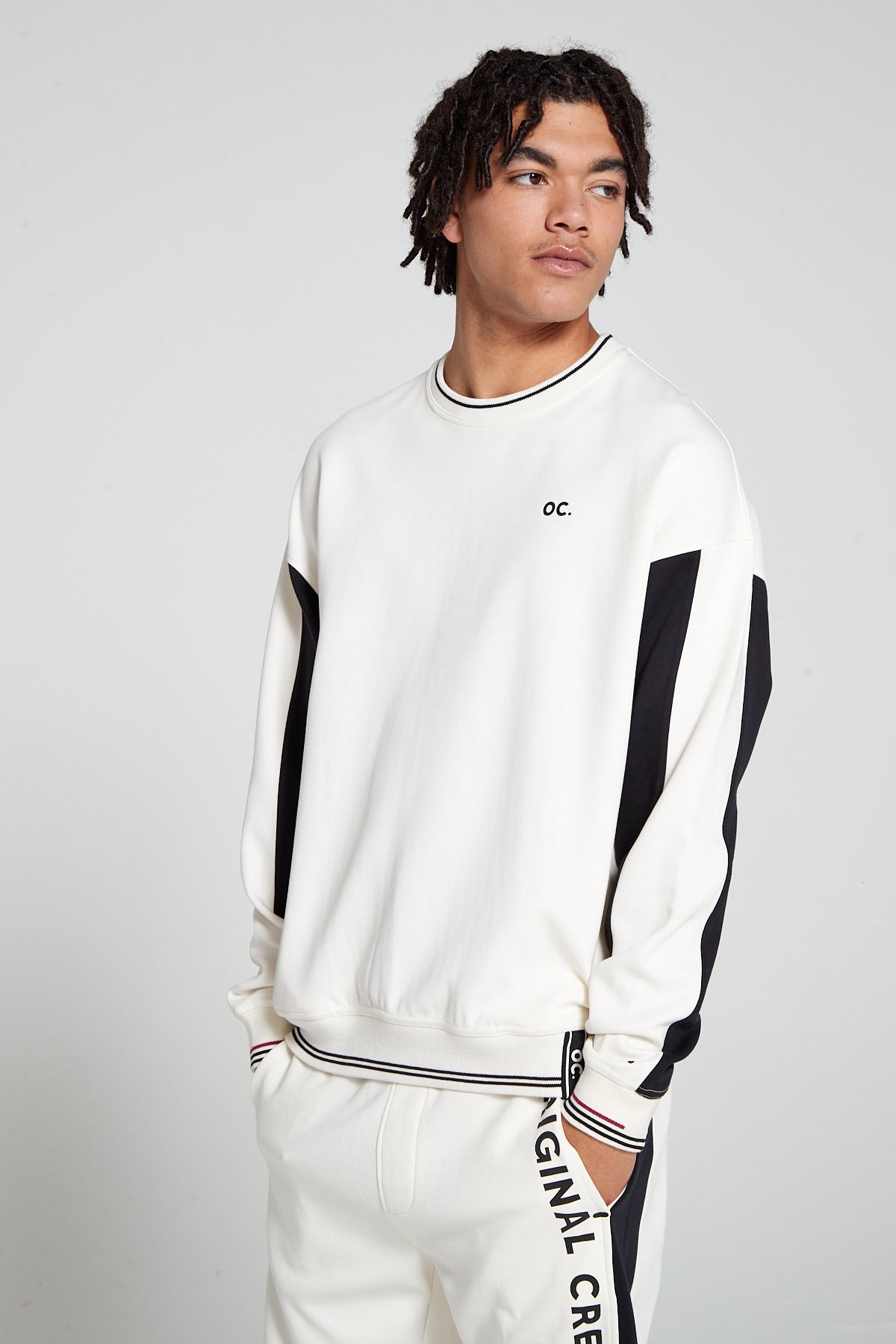 Front view of a model looking away wearing an Original Creator Retro Sweatshirt in Off White color. Showing the color combination of white and black. Sweatshirt features minimal OC branding on upper left side of chest with hidden pockets on the either side