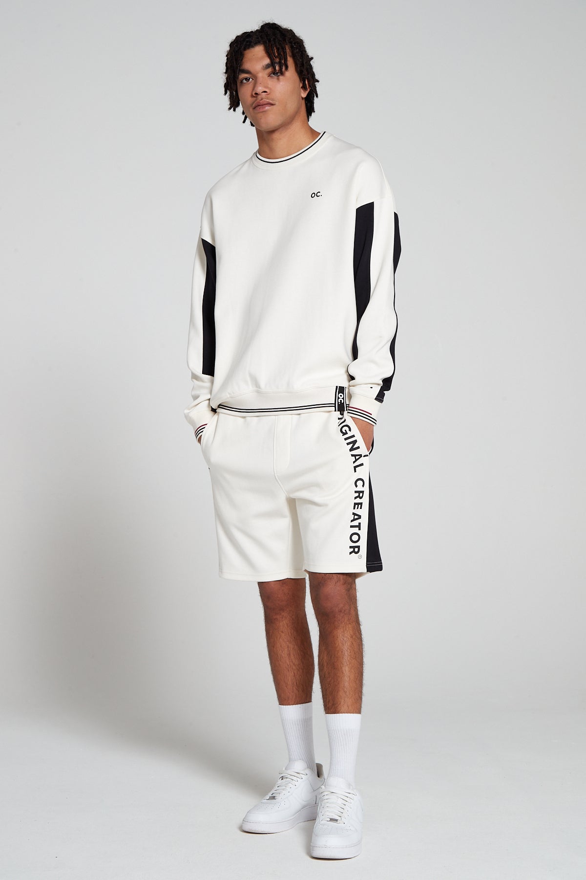 Full look of a model wearing Original Creator Off White zip pocket shorts with vertical branding on left thigh and featuring a matching Track set of Off white Sweatshirt and putting hands in the shorts pockets