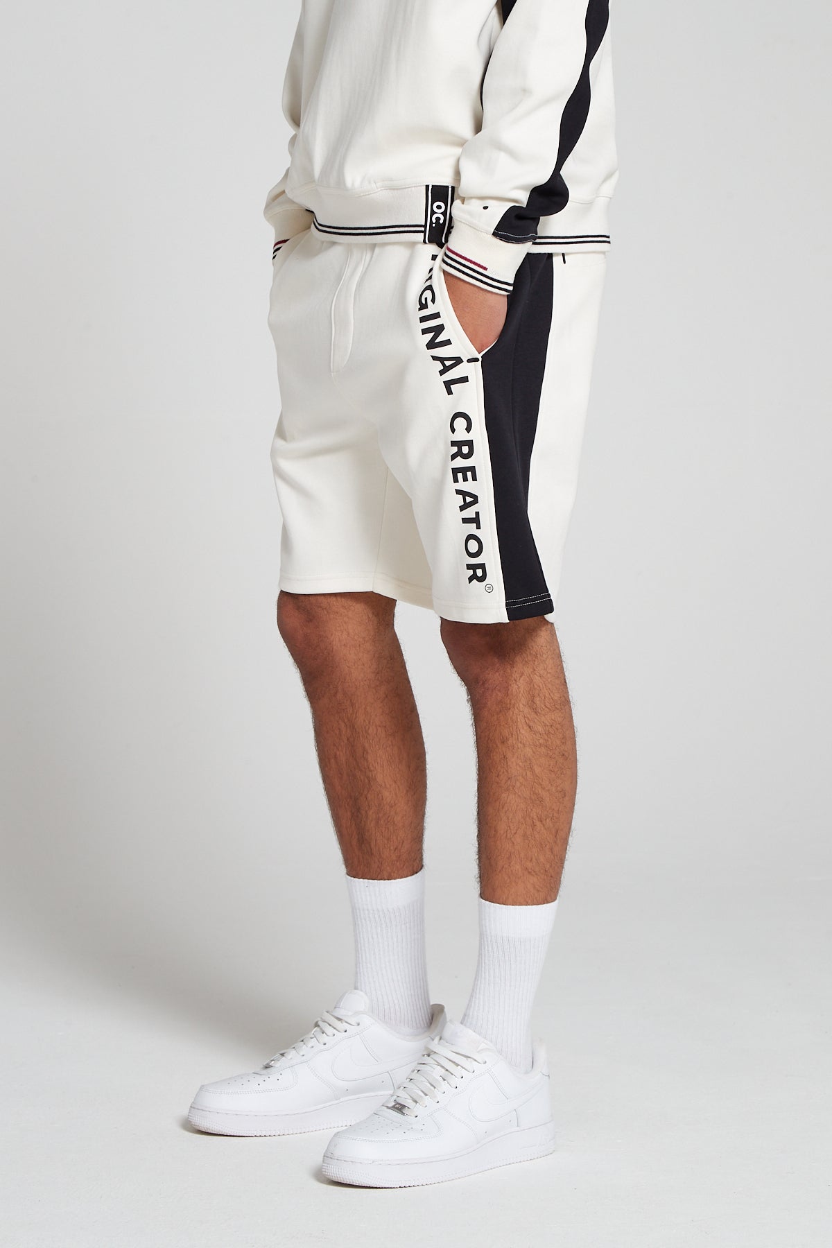 A bottom half side view of model wearing Original Creator Track 2.0 Retro Shorts in Off White Color. Model is showcasing the length of shorts and has hands in the pockets. Also highlighting the combination with off white long sleeve sweatshirt as well as vertical branding on left thigh.