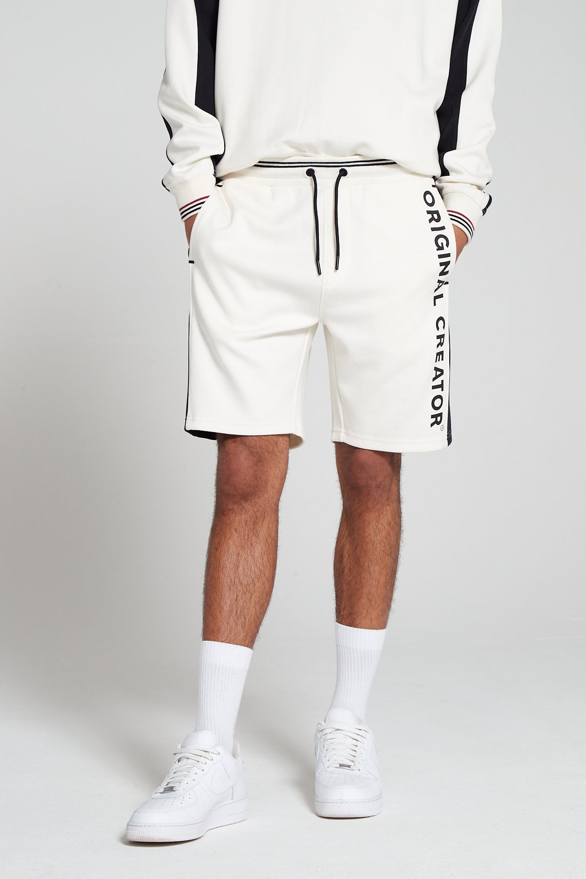 A bottom half front view of model wearing Original Creator Track 2.0 Retro Shorts in Off White Color. Model is showcasing the length of shorts and has hands in the pockets. Also highlighting the combination with off white long sleeve sweatshirt as well as vertical branding on left thigh.  