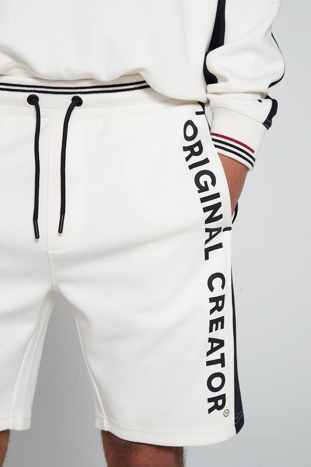 A close up shot of Original Creator Track 2.0 Retro Shorts in Off White Colour. Model is showcasing the vertical Original Creator branding and wearing a same colour track set. Model has his hands in the shorts pockets showcasing the shirt sleeves 