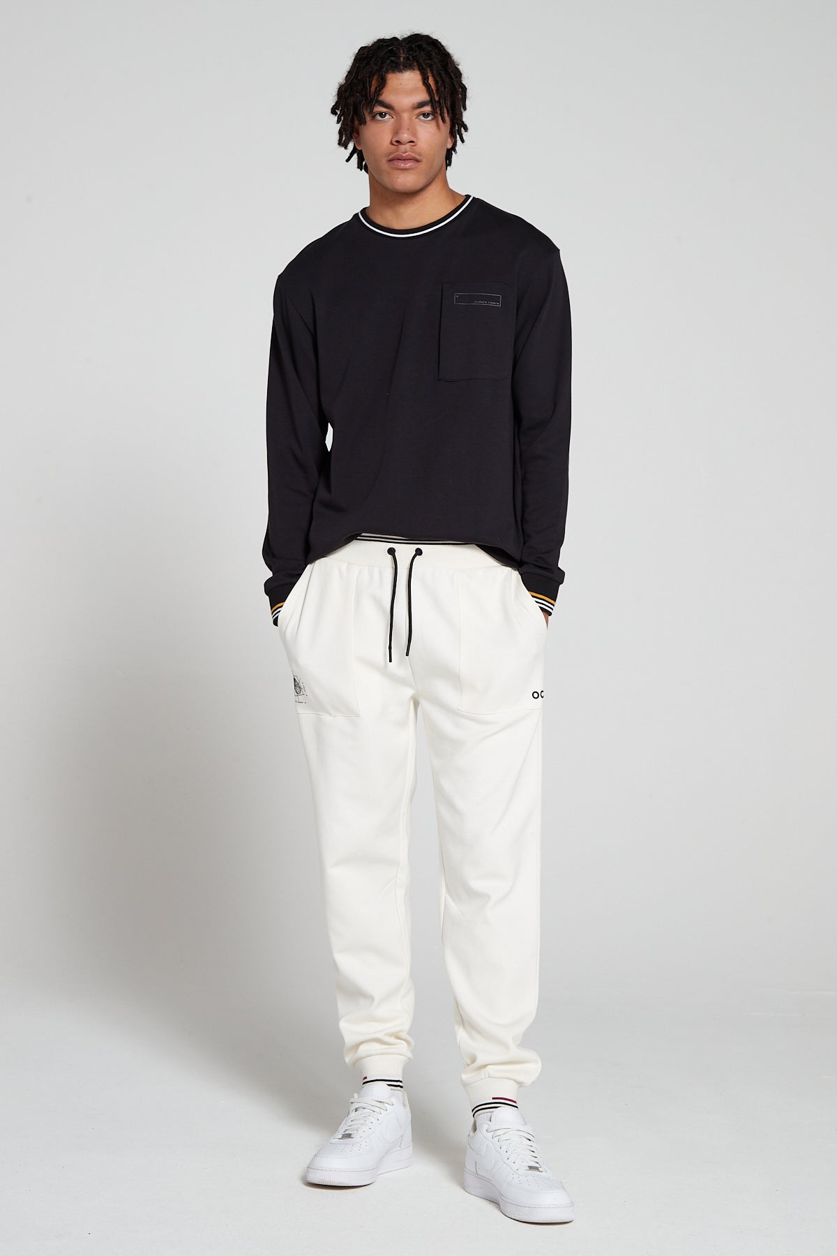 Full look of a model wearing Original creator Zip Pocket Long Sleeve Jet black T shirt with very low branding and features water repellent technology zip pocket and wearing Original creator Off White Classic joggers and shirt tucked in from the front