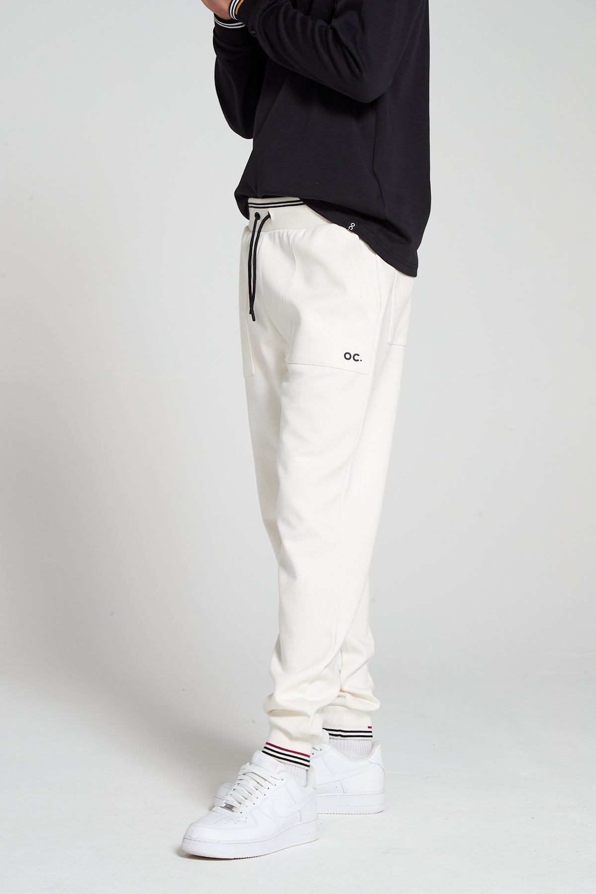 A bottom half shot of model wearing Original Creator Track 2.0 Classic Joggers in Off white Colour. Model is showcasing the length of Joggers and branding on the left thigh. Also highlighting the combination with Black long sleeve T shirt. 