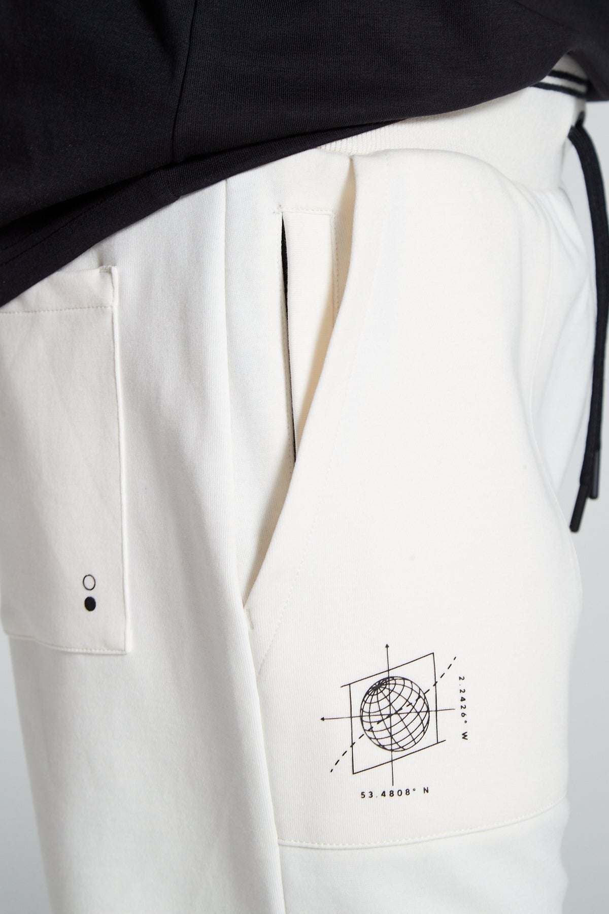 A side angle and close up shot of Original CreatorOff white Classic Joggers with a total of four side pockets includes two zip pockets. Close up shot also highlights a back pocket and globe branding under the pocket on right side.