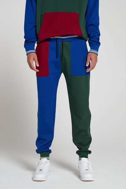 A bottom half shot of model wearing Original Creator Track 2.0 Classic Joggers in Multi color pattern with one leg in green and the other leg in blue color. Model is highlighting the combination with a multi color hoodie to complete the track set