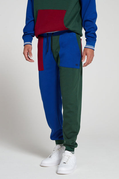A bottom half shot of model wearing Original Creator Track 2.0 Classic Joggers in Multi color pattern with one leg in green and the other leg in blue color. Model is highlighting the combination with a multi color hoodie to complete the track set 