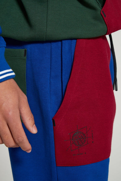 A close up angle of right leg of Original Creator Classic Joggers in Multicolor pattern includes a zip pocket and regular pocket. Close up shot also highlights globe branding under the red pocket on right side. Also includes a green pocket at the back on blue colored leg of jogger