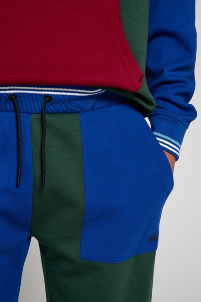 A close up angle of green colored left leg of Original Creator Classic Joggers in multicolor pattern includes a zip pocket and regular pocket. Close up shot also highlights OC branding under the blue pocket on green colored leg on left side.